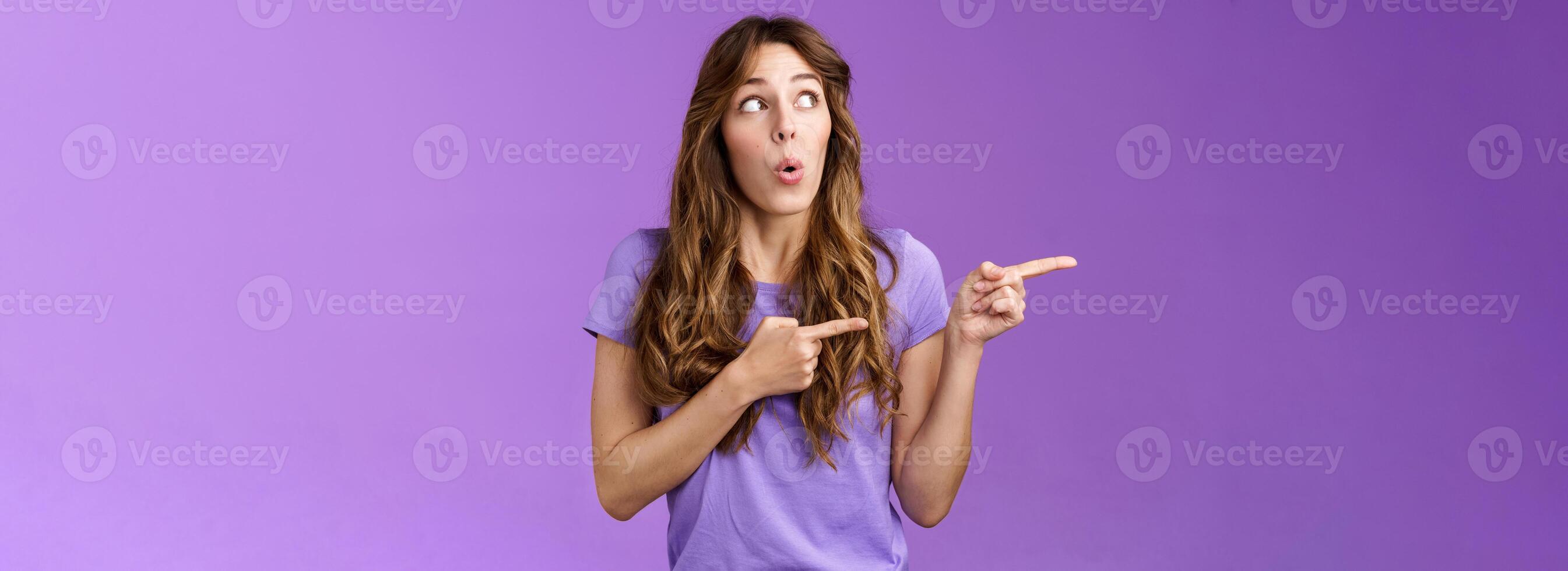 Curious surprised cute curly-haired caucasian female coworker discuss project amazed awesome cool result turn pointing left impressed folding lips wow tempating try new product purple background photo