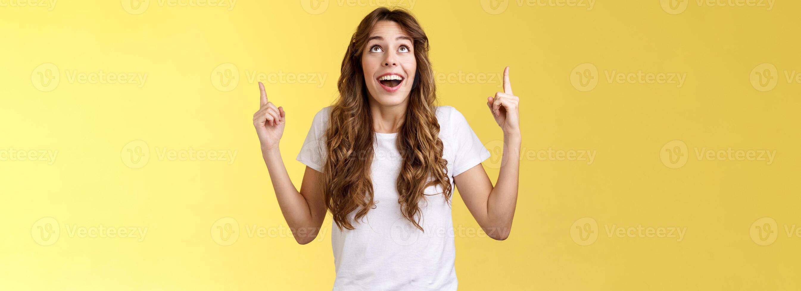 Happy impressed attractive girl long curly haircut drop jaw amused smiling surprised delighted look pointing up index fingers stare admiration fascinated awesome promo offer yellow background photo