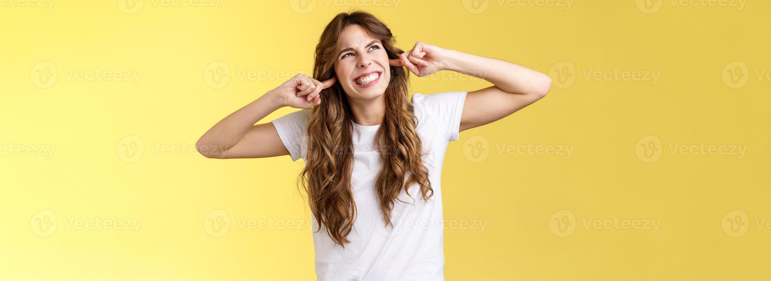 Angry displeased annoyed curly-haired beautiful girl grimacing make wry face stare up irritated noise comming from neighbours above clench teeth plug index fingers ears hate loud music playing photo