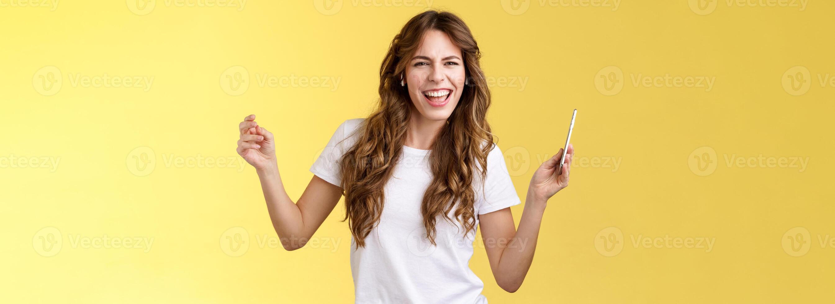 Sassy carefree joyful attractive woman having fun partying listen cool new song favorite artist dancing raise hands up squinting cheeky look camera lip sync hold smartphone yellow background photo