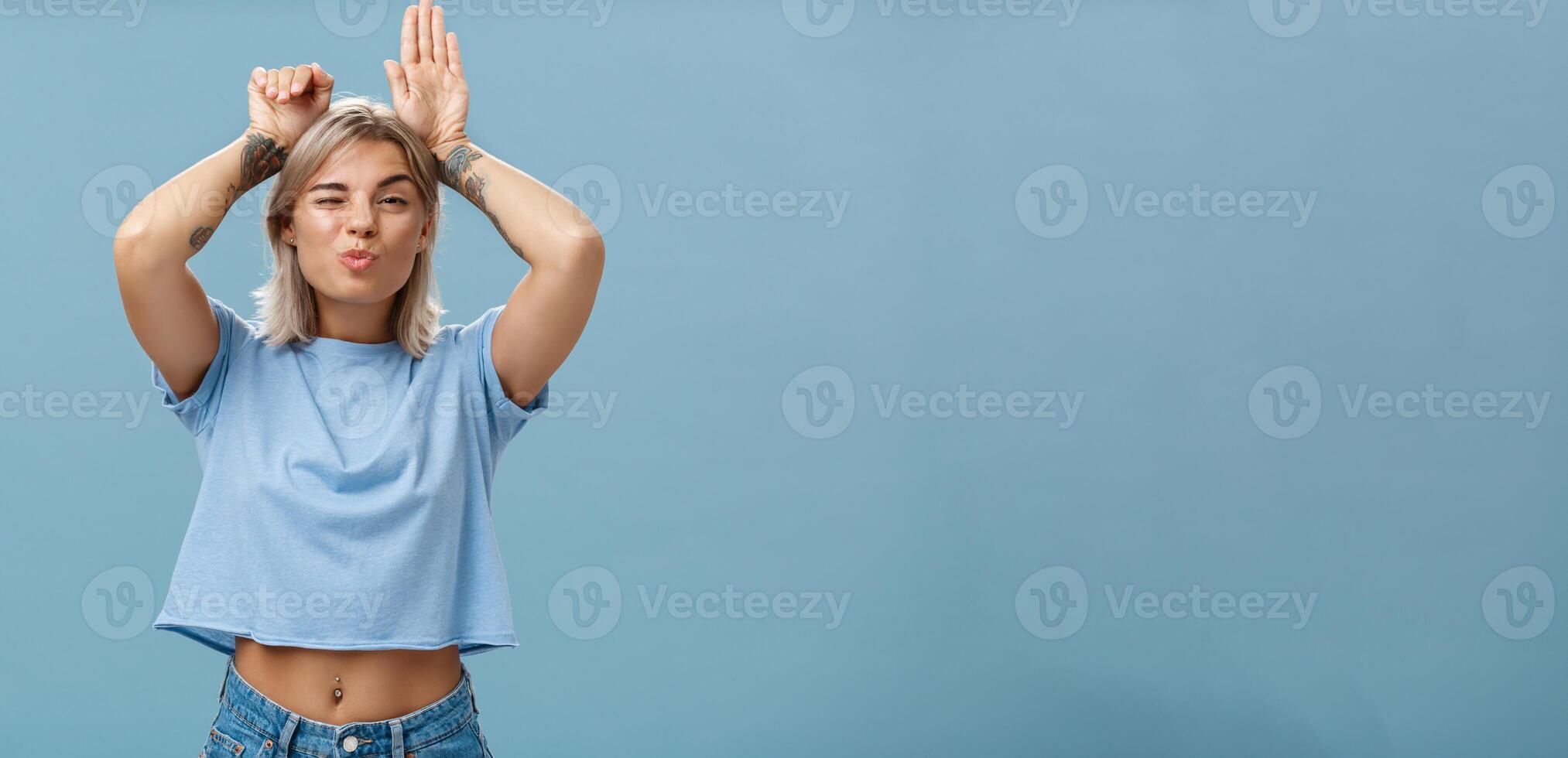Flirty attractive and stylish cool girlfriend with tattooed arms and pierced belly winking folding lips in air kiss showing bunny ears with palms on head standing over blue background playful photo