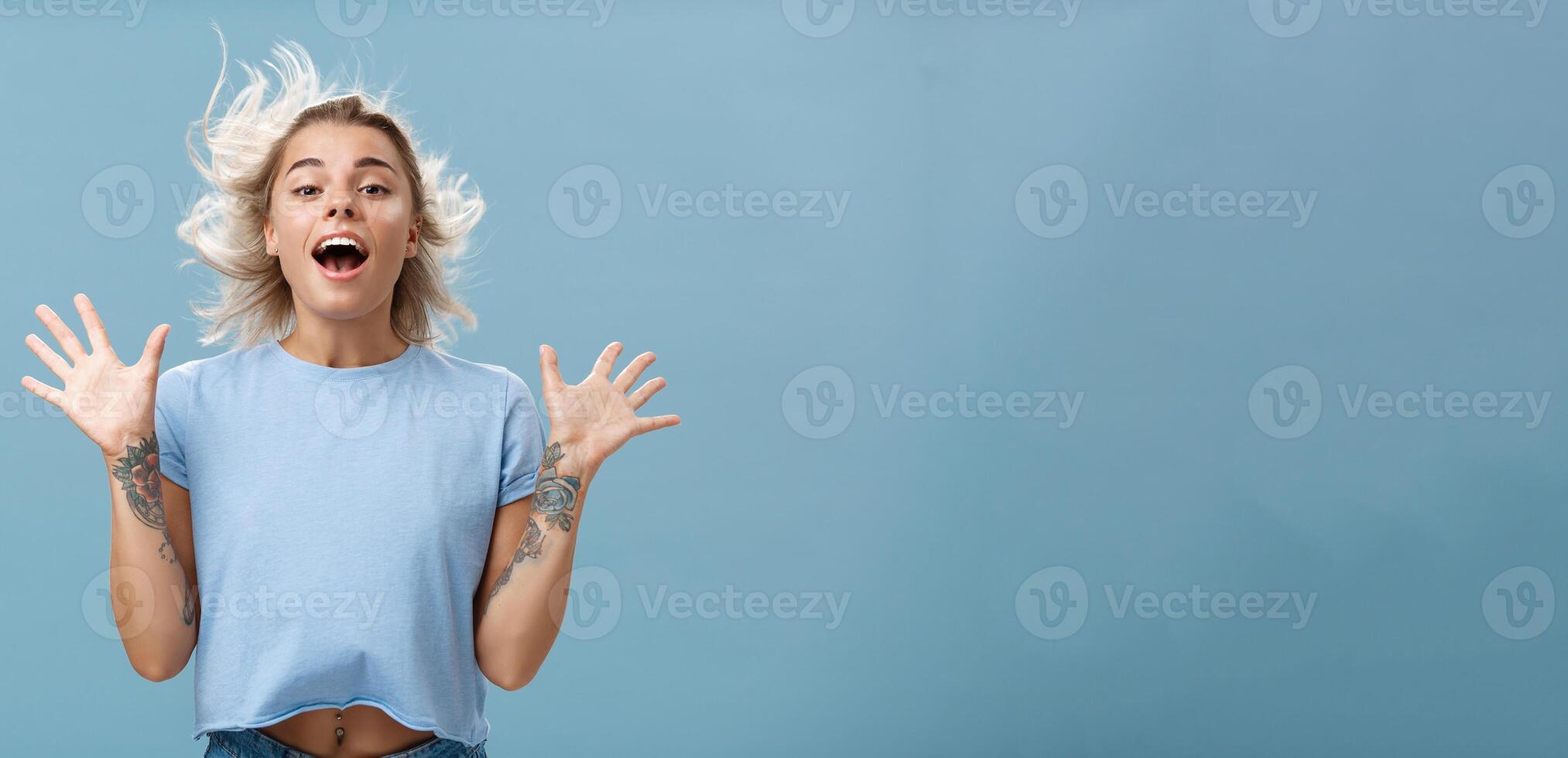Creative happy and playful beautiful blond girl with tattoos on arms raising palms up opening mouth facing wind while hair strands flicking on air jumping having fun over blue background photo