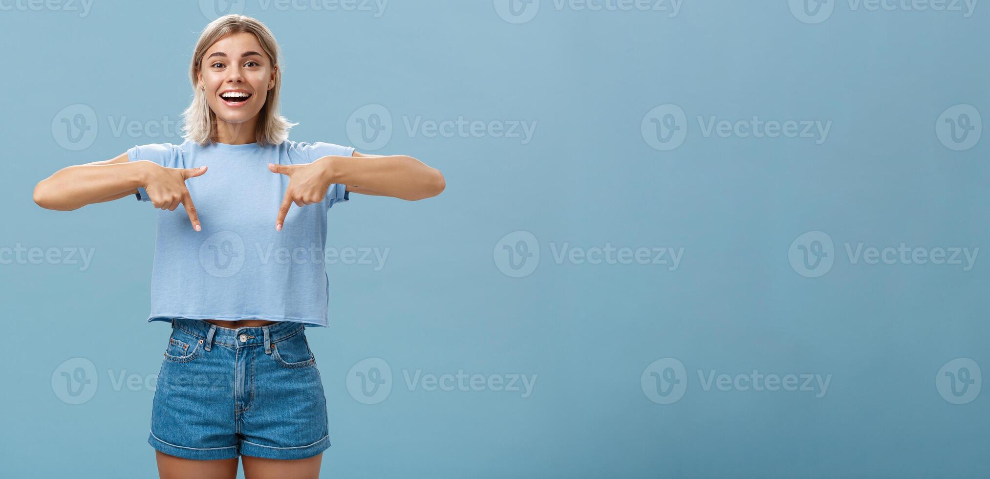 You have to see this. Portrait of joyful amused and happy stylish blonde female in trendy t-shirt pointing down with arms near chest smiling broadly showing amazing copy space over blue background photo