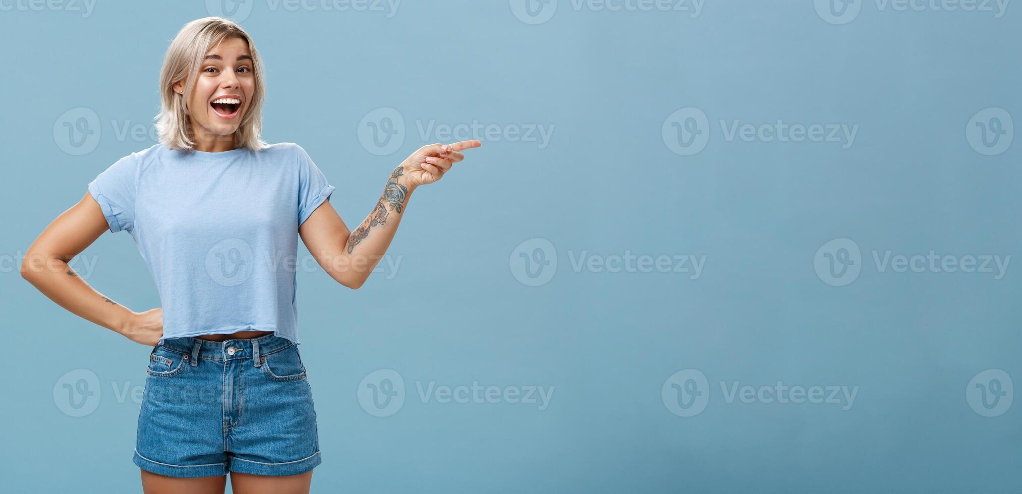 Studio shot of impressed and surprised entertained blonde female in summer outfit holding hand on waist pointing right and smiling joyfully being amazed and amused over blue background photo