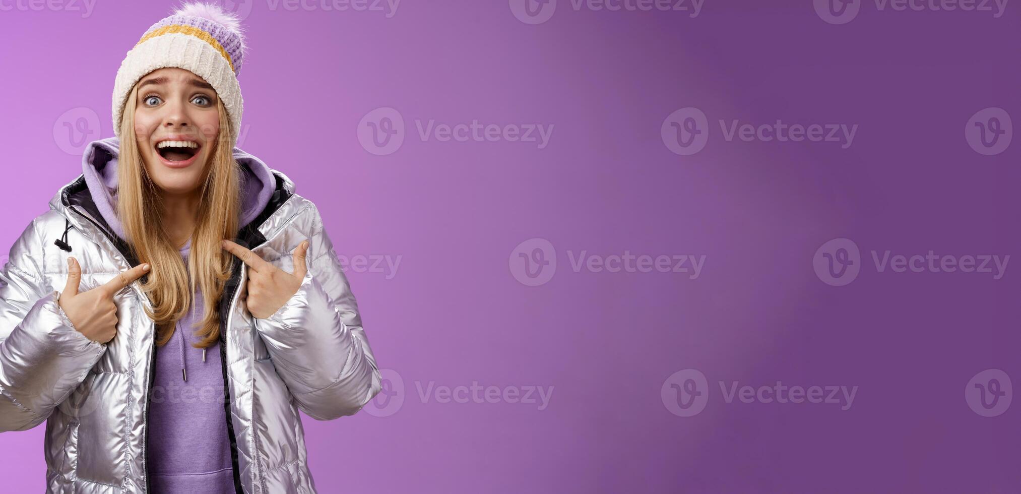 Surprised happy smiling joyful attractive woman pointing herself satisfied picked was chosen winner standing excited in silver shiny jacket winter hat cannot believe own luck, purple background photo