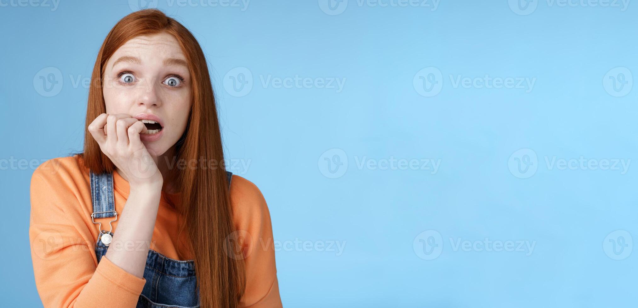 Scared unconfident anxious young trembling redhead girl wide eyes staring intense emotional biting fingernails, fan worry favorite character tv series dies standing nervously blue background photo