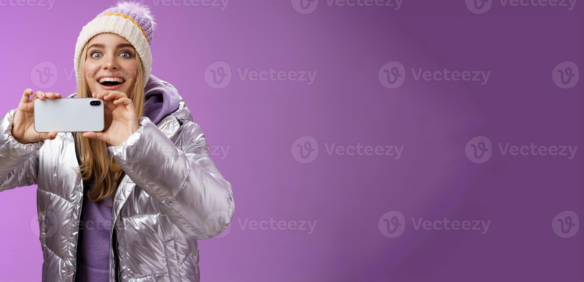 Amused fascinated female in silver jacket head smiling astonished excited look forward holding smartphone recording video taking shots famous person mobile phone camera, purple background photo