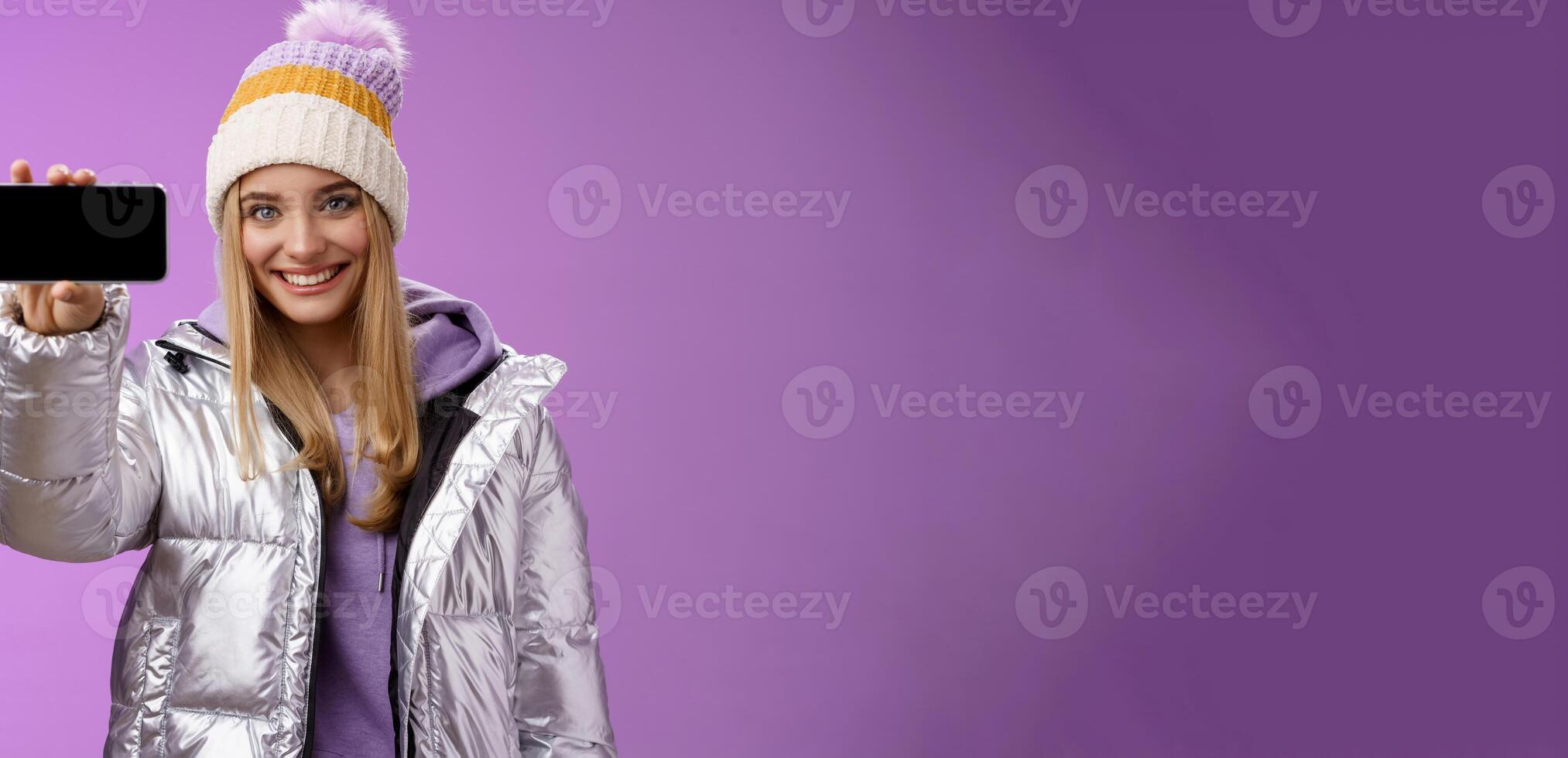 Confident friendly good-looking blond girl in outdoor silver glittering jacket hat hold smartphone horizontal showing mobile phone display assertive smile recommend use app, purple background photo