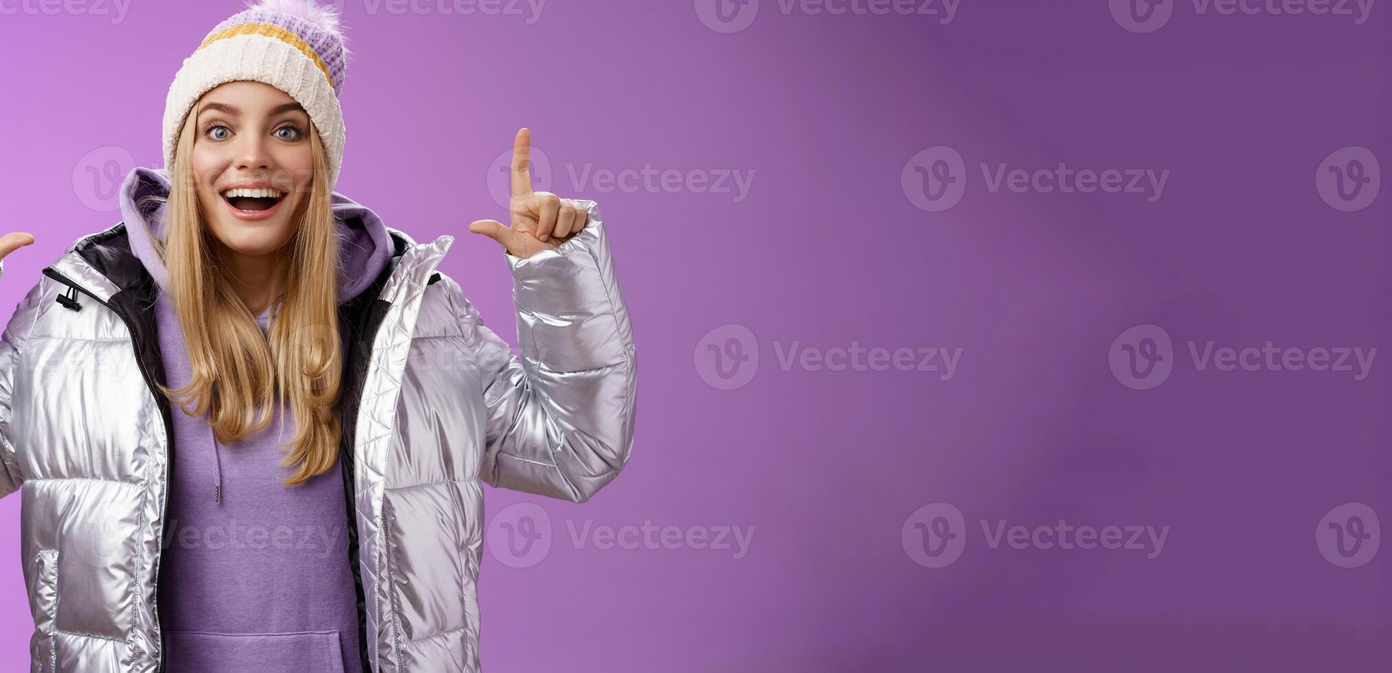 Excited carefree cheerful fair-haired european girl in silver jacket winter hat raising hands pointing up have excellent idea smiling broadly speaking passionately standing purple background photo