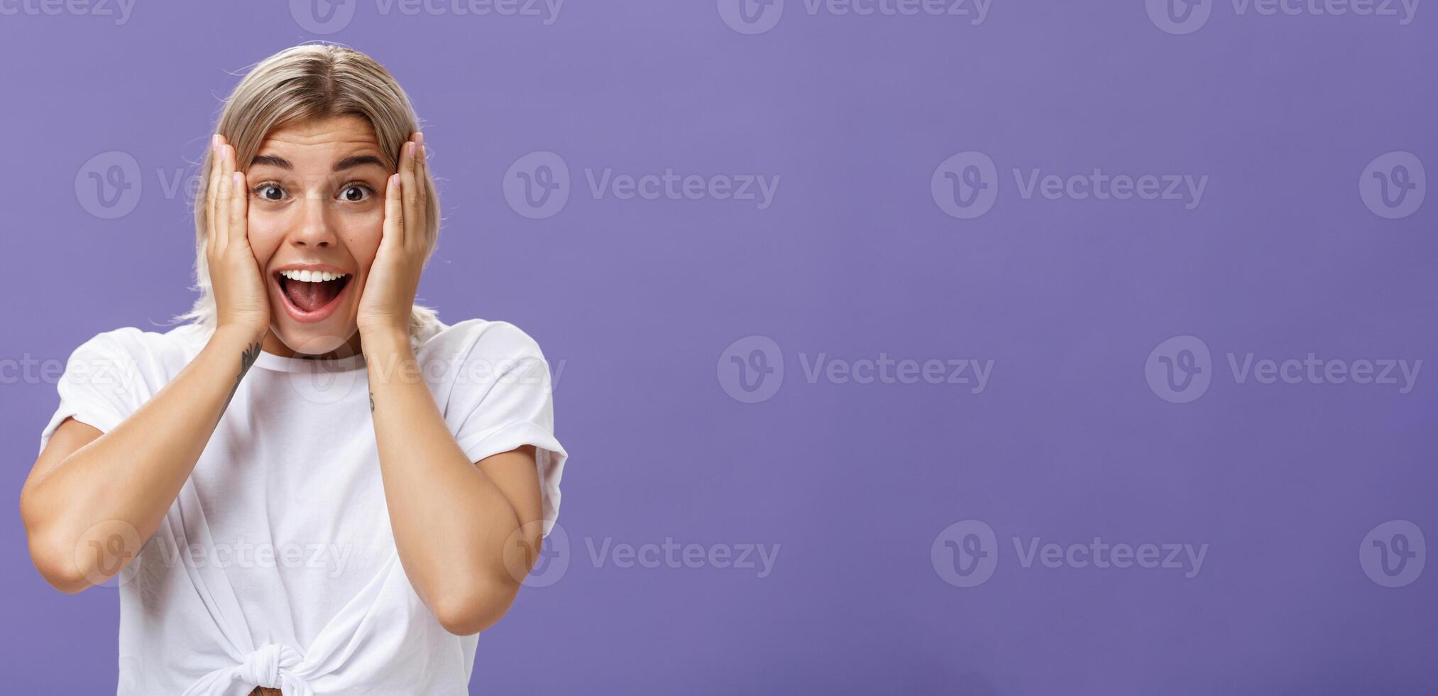 Amazed and thrilled happy european woman with fair hair clean skin and white teeth smiling joyfully holding hands on cheeks being impressed and amused with awesome news over purple wall photo