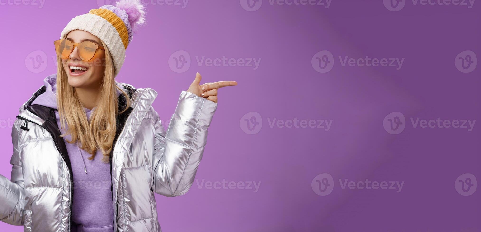 Cool sassy friendly-looking blond girl pointing different sides right left choose which way go look delighted carefree enjoy awesome winter vacation wear stylish silver jacket, hat sunglasses photo