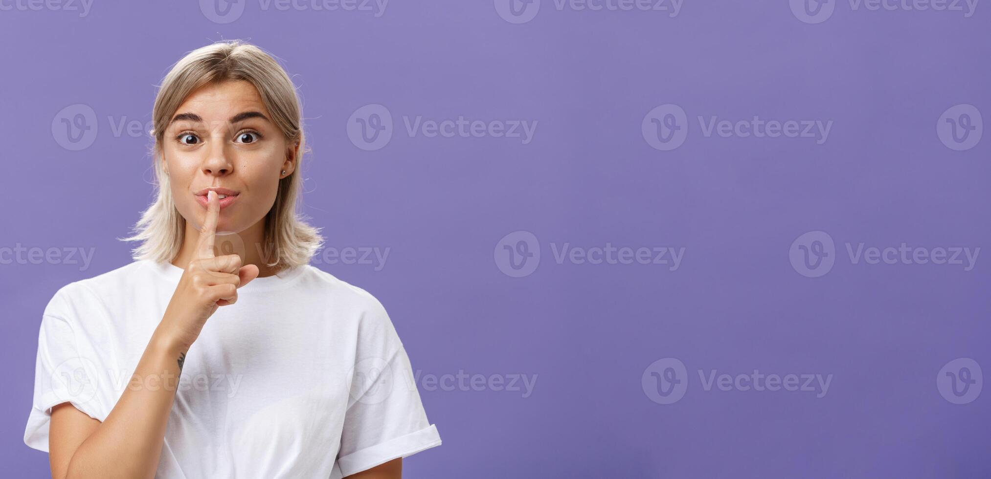 Charming creative and stylish beautiful tanned girl with fair hair shushing at camera with delighted thrilled look making up great idea wanting hide it and keep secret posing over purple wall photo