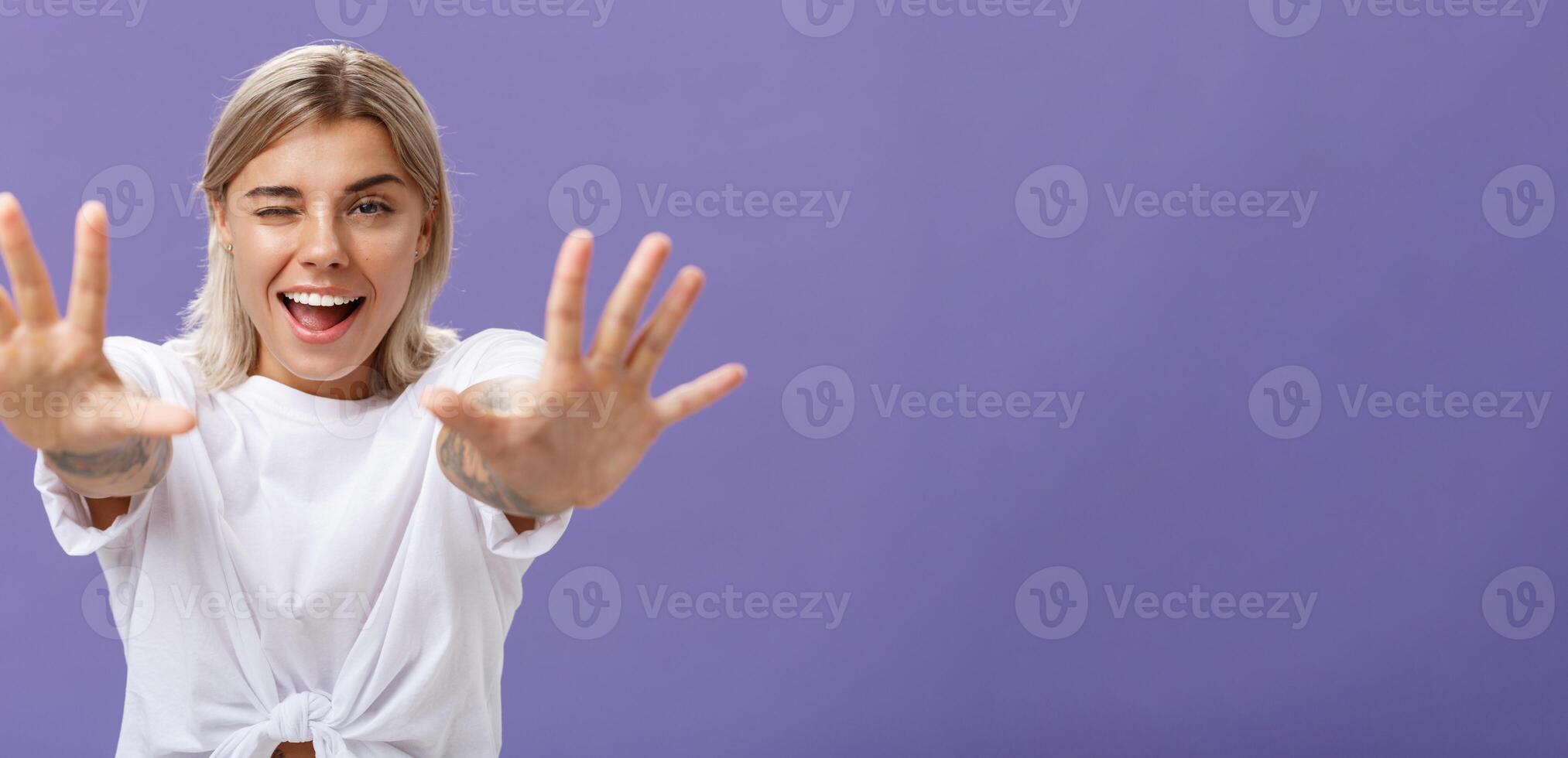 Reaching to you. Portrait of flirty good-looking and confident stylish woman with tattoos on arms winking sticking out tongue playfully and smiling pulling hands towards camera over purple background photo