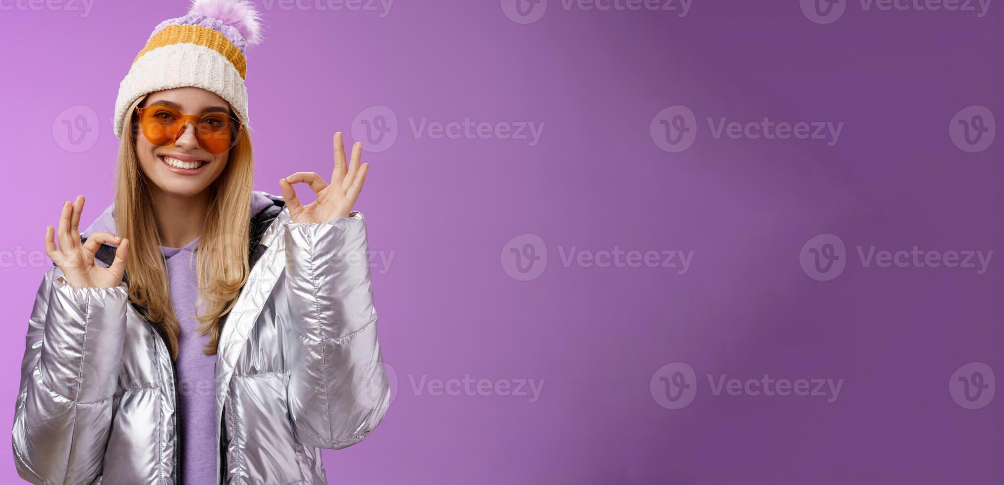 Everything fine thanks. Charming flirty blond confident woman in silver stylish jacket sunglasses winter hat show okay no problem ok gesture smiling affirmative, liking awesome day purple background photo