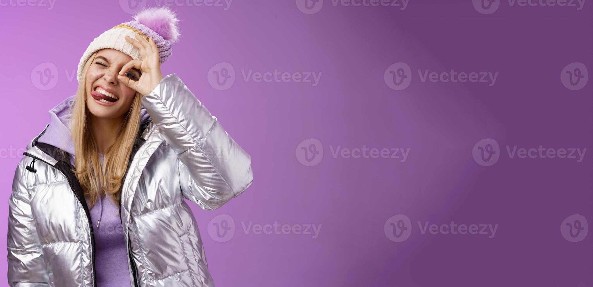 Friendly joyful caucasian blond girl in hat warm stylish shiny silver jacket tilt head happily show tongue smiling broadly enjoying awesome ski resort vacation travelling winter, purple background photo