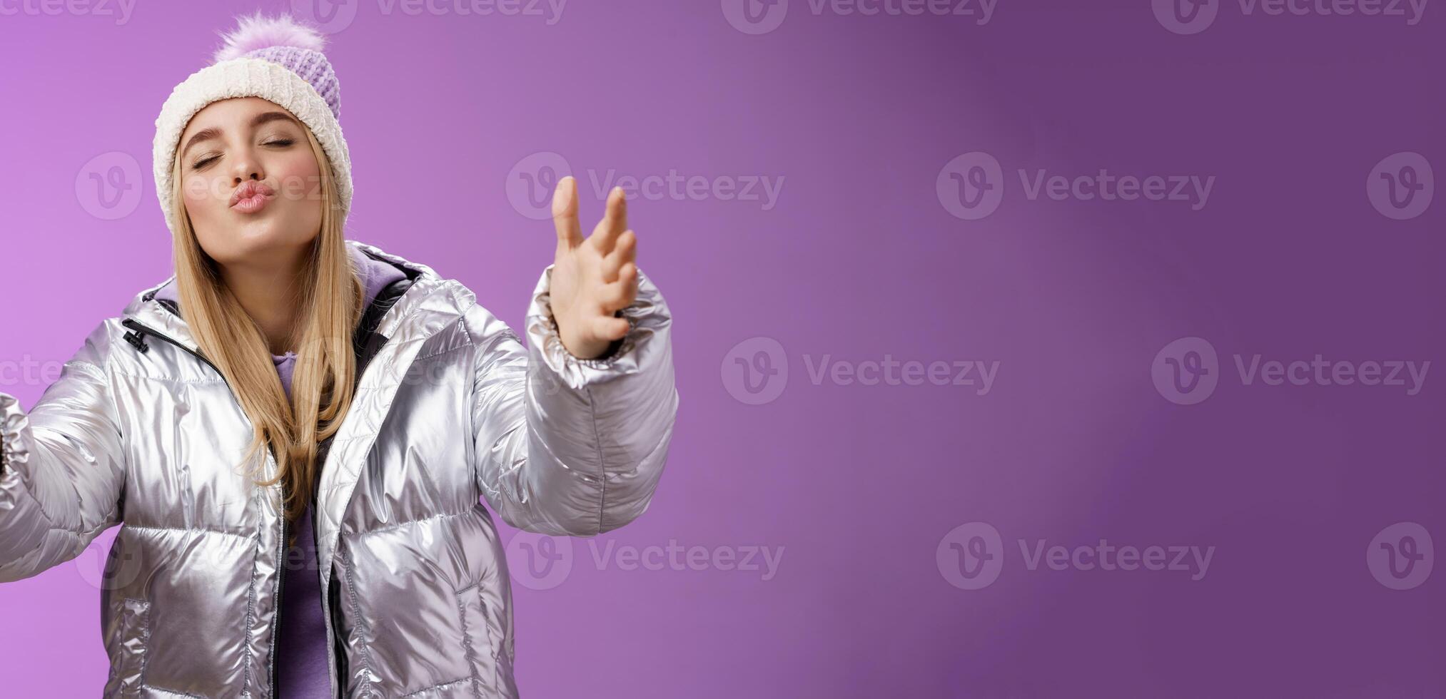 Romantic charming caucasian blond woman in cute hat silver stylish jacket pulling arms forward wanna embrace tight cuddle boyfriend folding lips close eyes give kiss, standing purple background photo