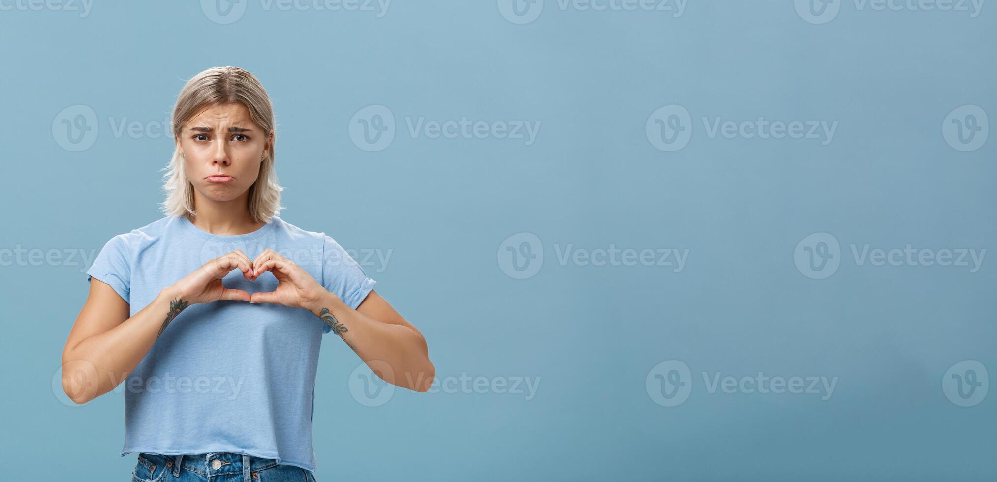 Heart being broken. Sad and gloomy heartbroken girl with blond hair tattoos on arms and tanned skin pursing lips whining and complaining making love sign over breast standing unhappy near blue wall photo