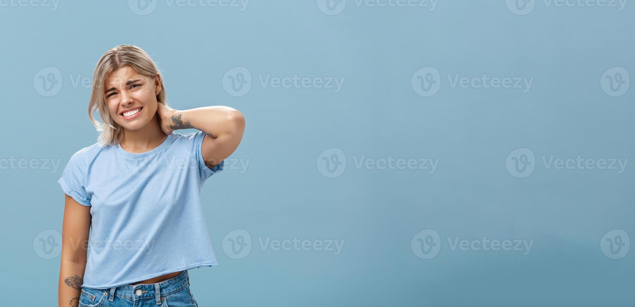 Girl unwilling to go with friend feeling awkward and unsure how say no frowning and clenching teeth making apologizing face rubbing neck behind saying sorry while rejecting offer over blue background photo