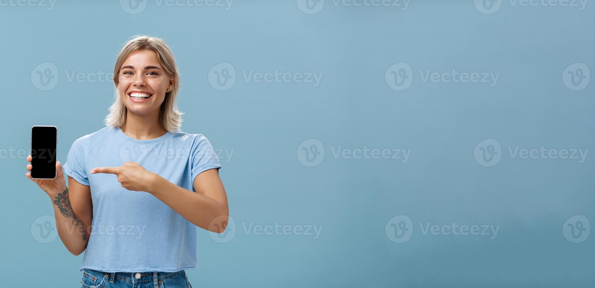 Cute enrgized girl showing smartphone. Charming blonde young female with cool tattoos laughing and smiling from happiness and joy pointing at device holding phone with screen faced to camera photo