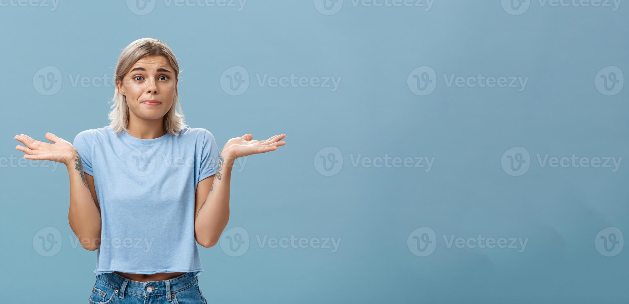 No idea how help sorry. Portrait of unsure gloomy girlfriend feeling regret shrugging with hands near shoulders frowning and pursing lips being uncertain and unaware over blue background photo