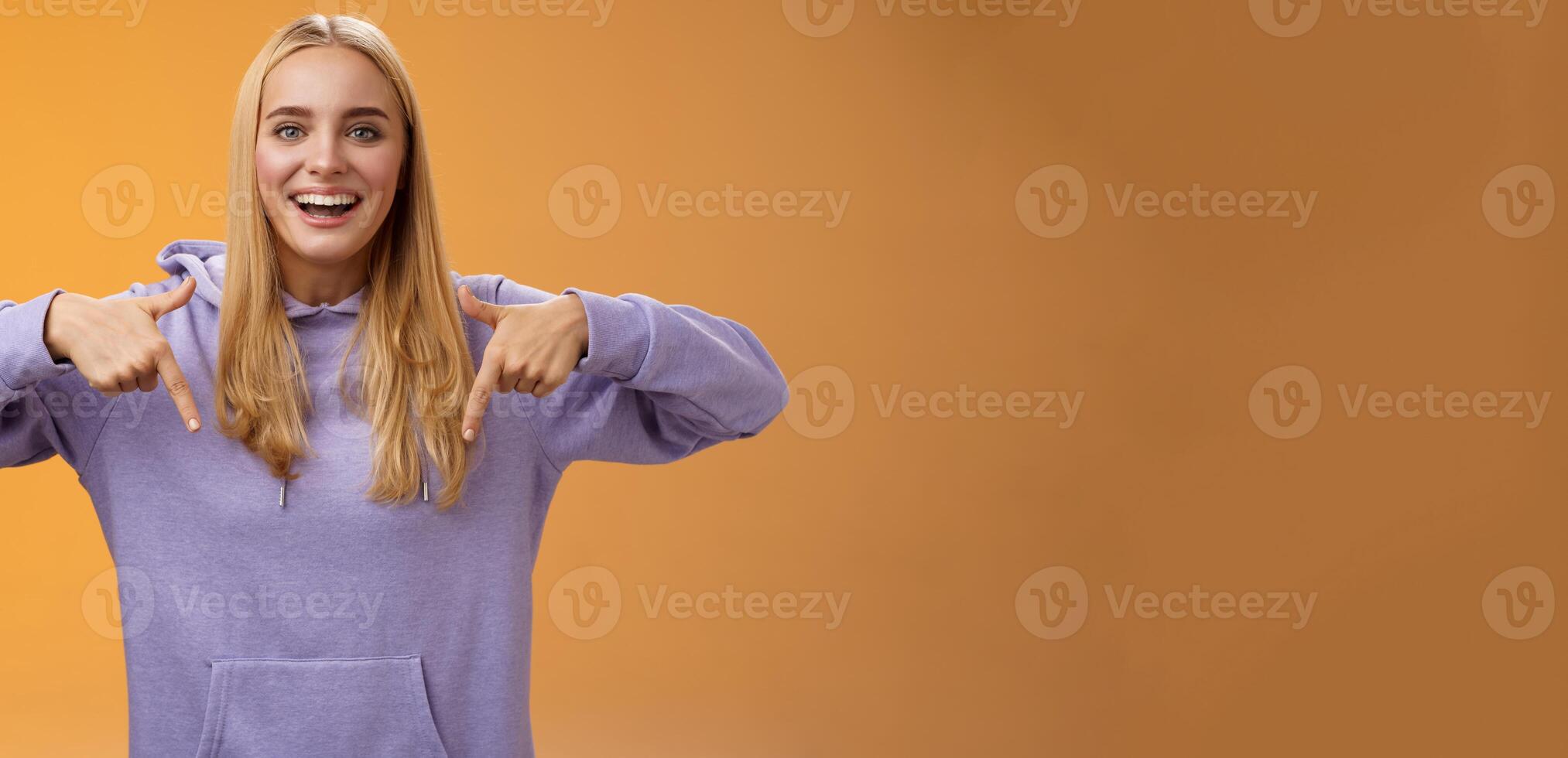 Amused joyful nice blond girlfriend pointing down present cool new product smiling broadly recommending try check out standing orange background happily grinning in hoodie photo