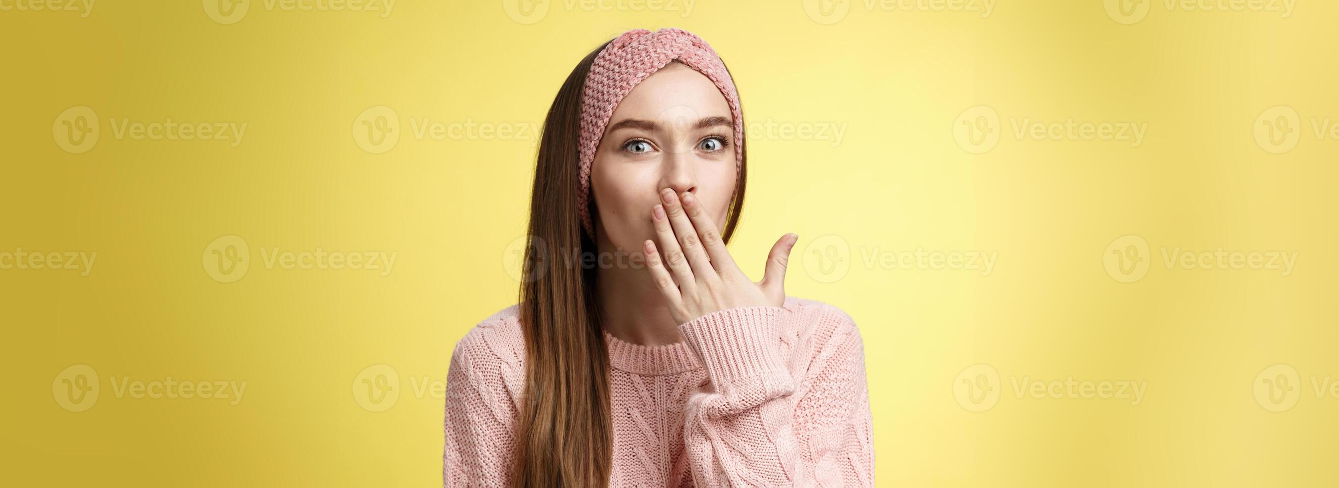 Talkative young cute office worker wearing sweater, knitted headband holding palm on mouth surprised and amazed, learning interesting shocking rumor, gossiping entertained over yellow wall photo