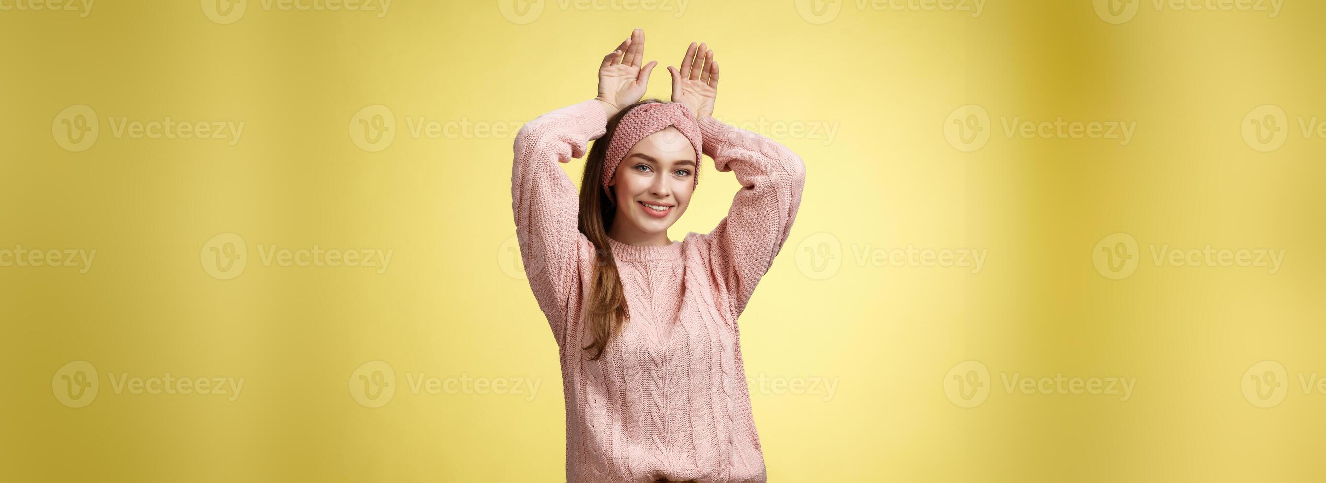 Nice charming playful young european female having fun, playing mimicking bunny showing rabbit ears with hands behind head, smiling cute, tender, happy greeting boyfriend welcome back ove yellow wall photo