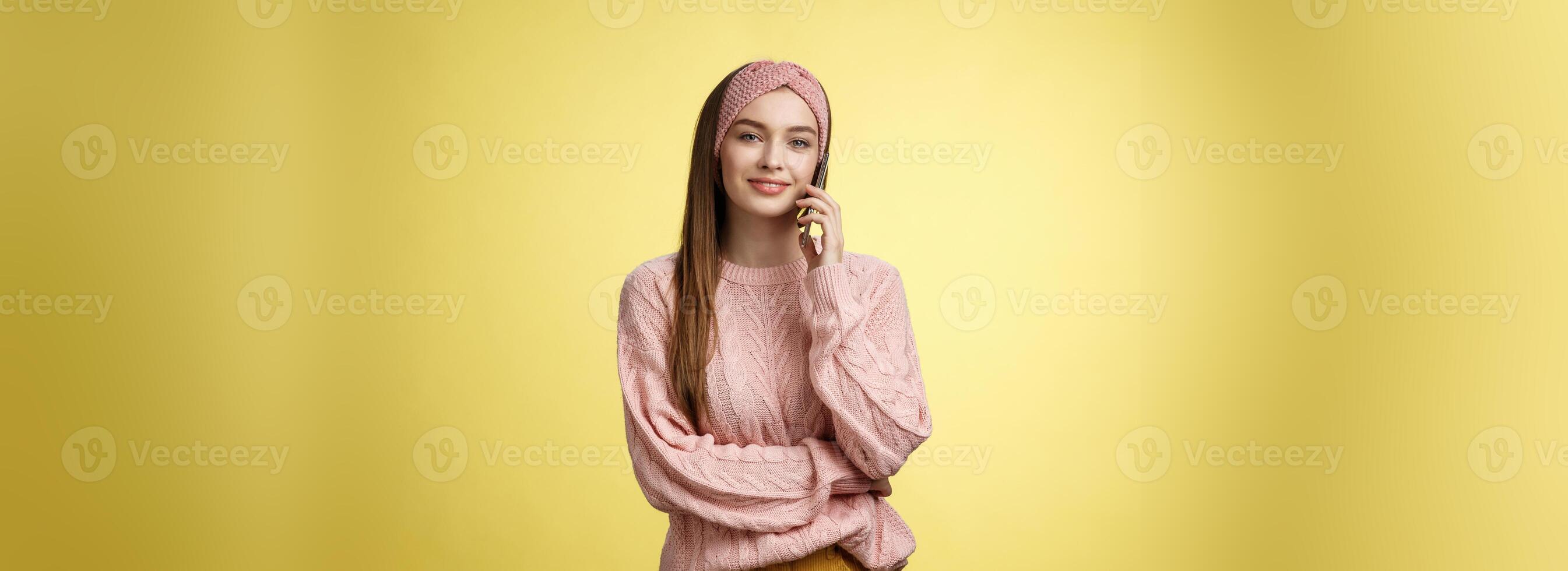 Stylish young girlfriend having pleasant conversation via phone holding smartphone near ear talking, making reservation in restaurant, ordering food delivery, smiling, posing against yellow wall photo