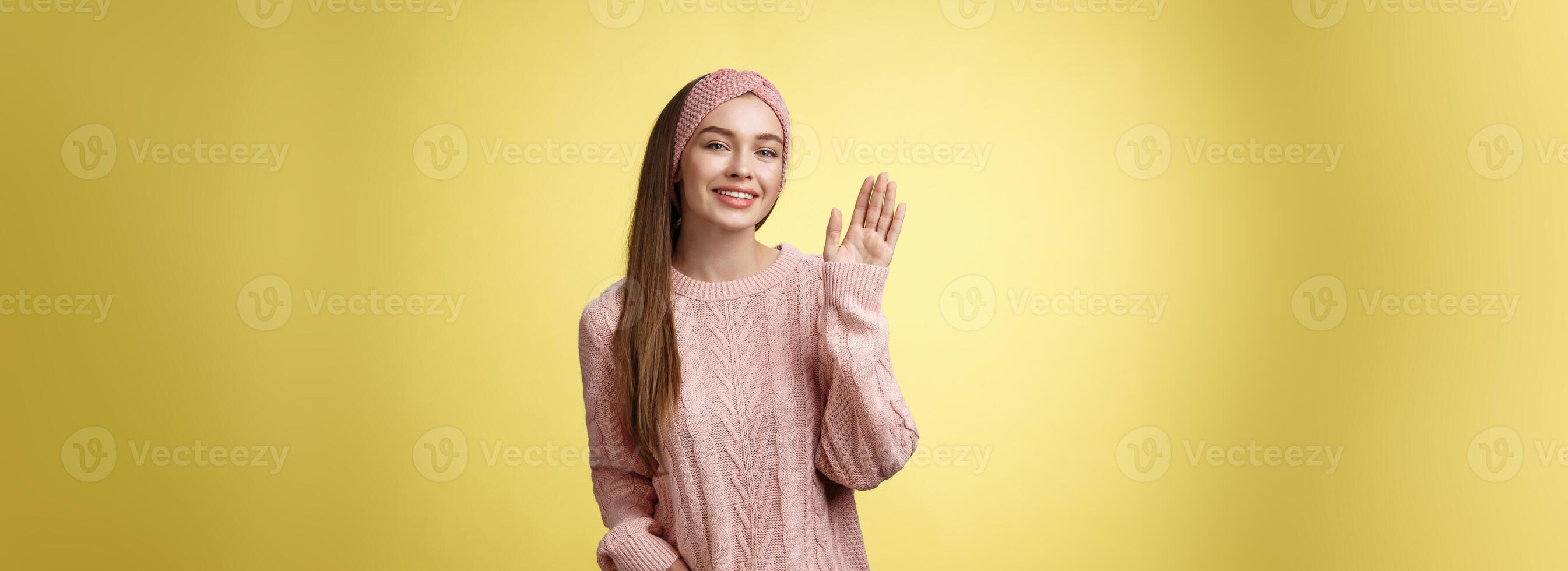 Hello adventures. Charming glamour 20s girl in sweater, knitted headband waving palm saying hi friendly smiling, holding hand in pocket gretting friends, posing positive and carefree over yellow wall photo