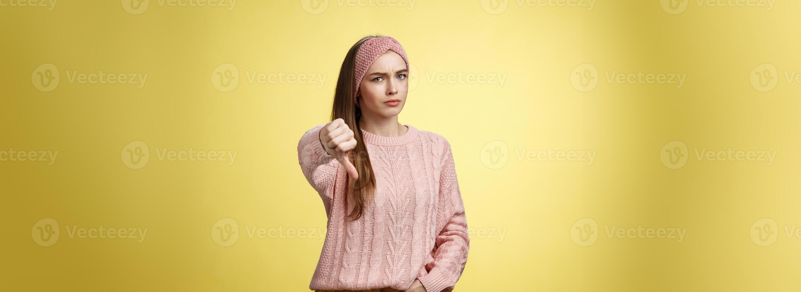 no Frío. frustrado disgustado y discrepar linda joven mujer vistiendo de punto suéter ceñudo demostración pulgar hacia abajo sonriendo crítico, expresando desaprobación de indispuesto situación terminado amarillo pared foto