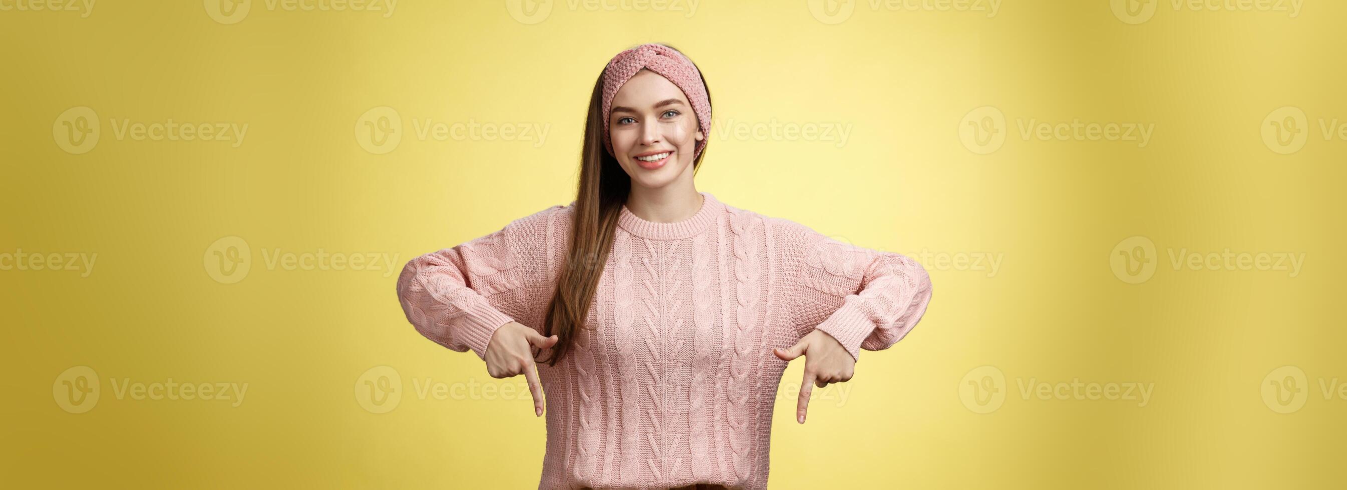 Tender young 20s european girl in sweater smiling broadly finally take-off braces pointing down promoting skillful dentist professional. Female student standing happy positive over yellow wall photo