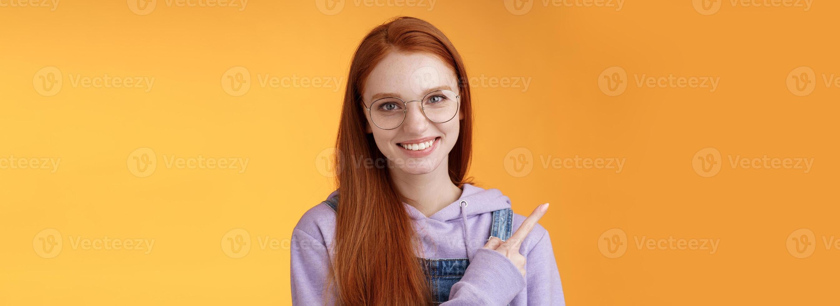 Friendly good-looking modern redhead young girl pointing left index finger showing awesome place suggesting go hang out smiling joyfully casually talking discuss new product, orange background photo