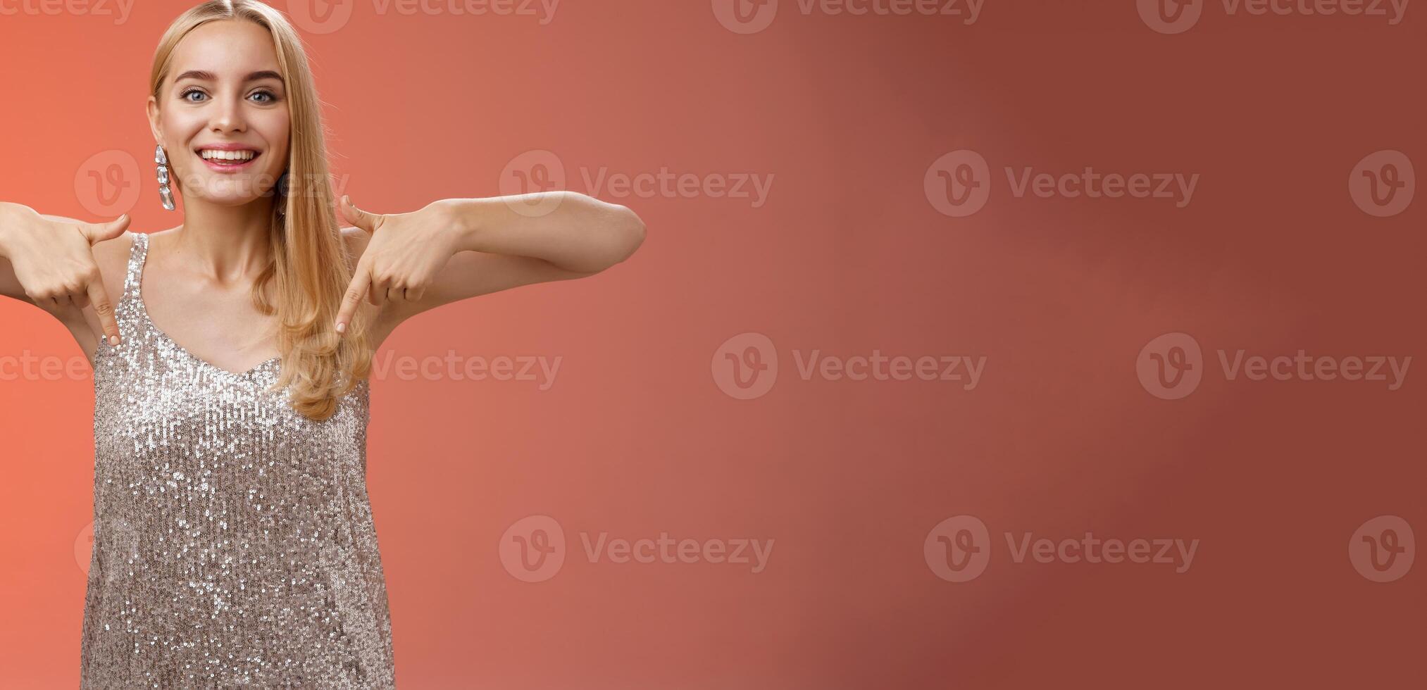 Attractive glamour blond woman in silver glittering dress pointing down smiling excited showing awesome party place hang-out inviting try-out standing pleased red background enjoying perfect evening photo