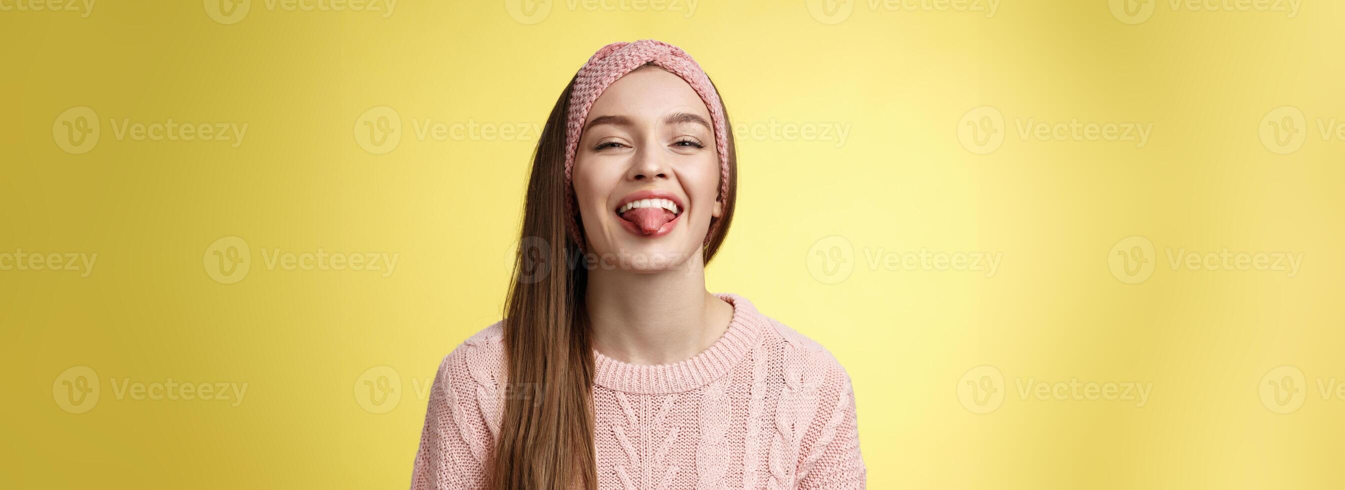 Positive entertained cute funny glamour young european girl in sweater, knitted trendy headband smiling fooling around showing tongue playfully, mocking friend enjoying sunny day over yellow wall photo