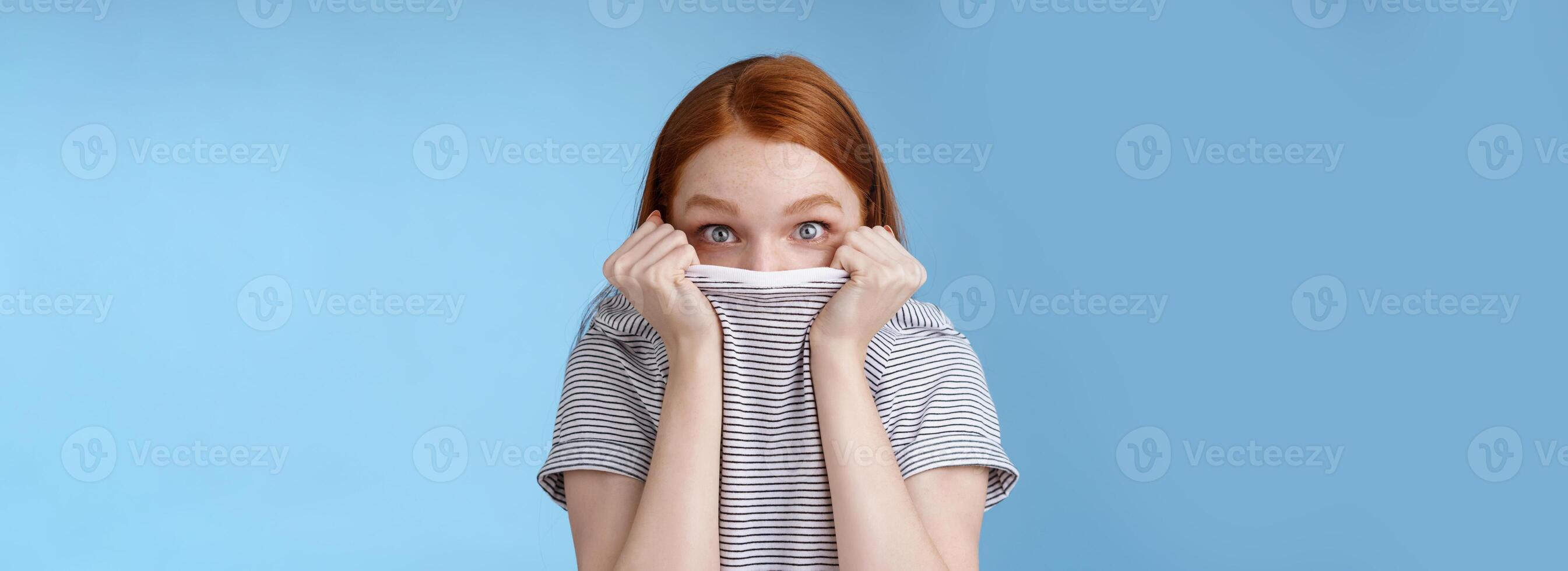 You cannot see me. Silly cute young teenage redhead girl hiding face pulling t-shirt head peeking look excited surprised camera hold collar standing funny under disguise, blue background photo