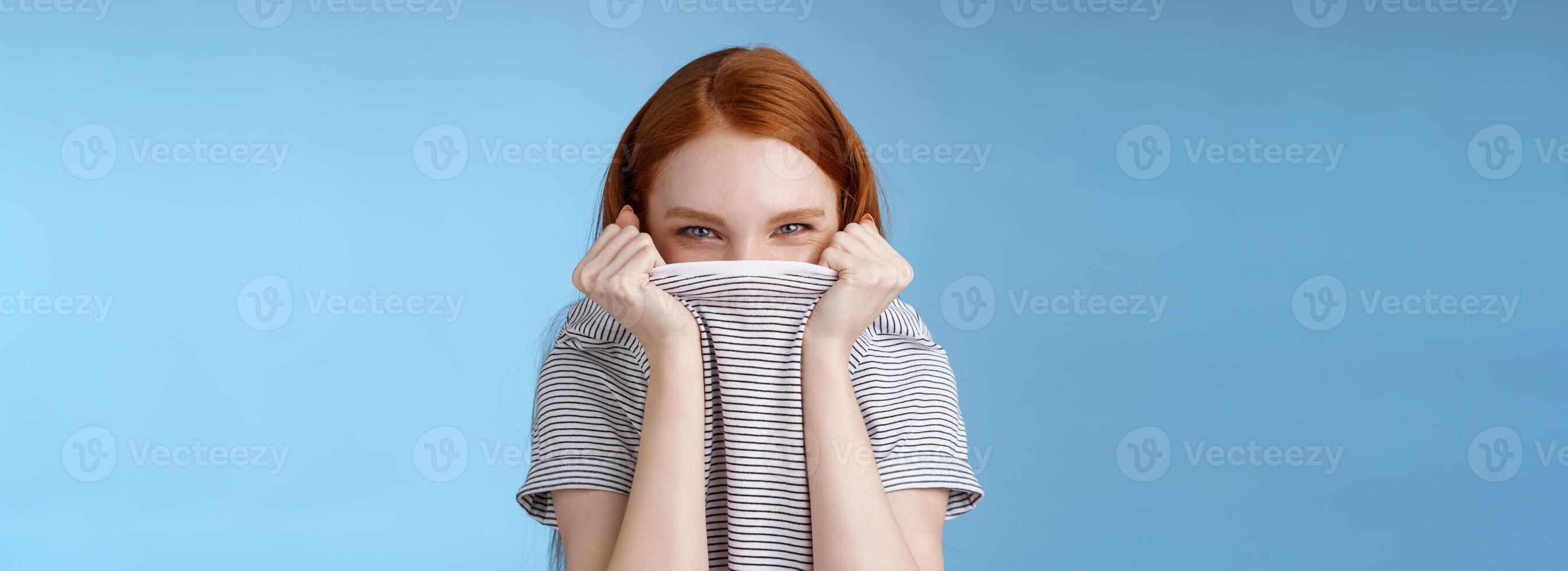 Silly flirty amused attractive playful redhead girlfriend hiding face pulling t-shirt head squinting devious mysteriously giggle laughing hope disguise pranking friend standing blue background photo