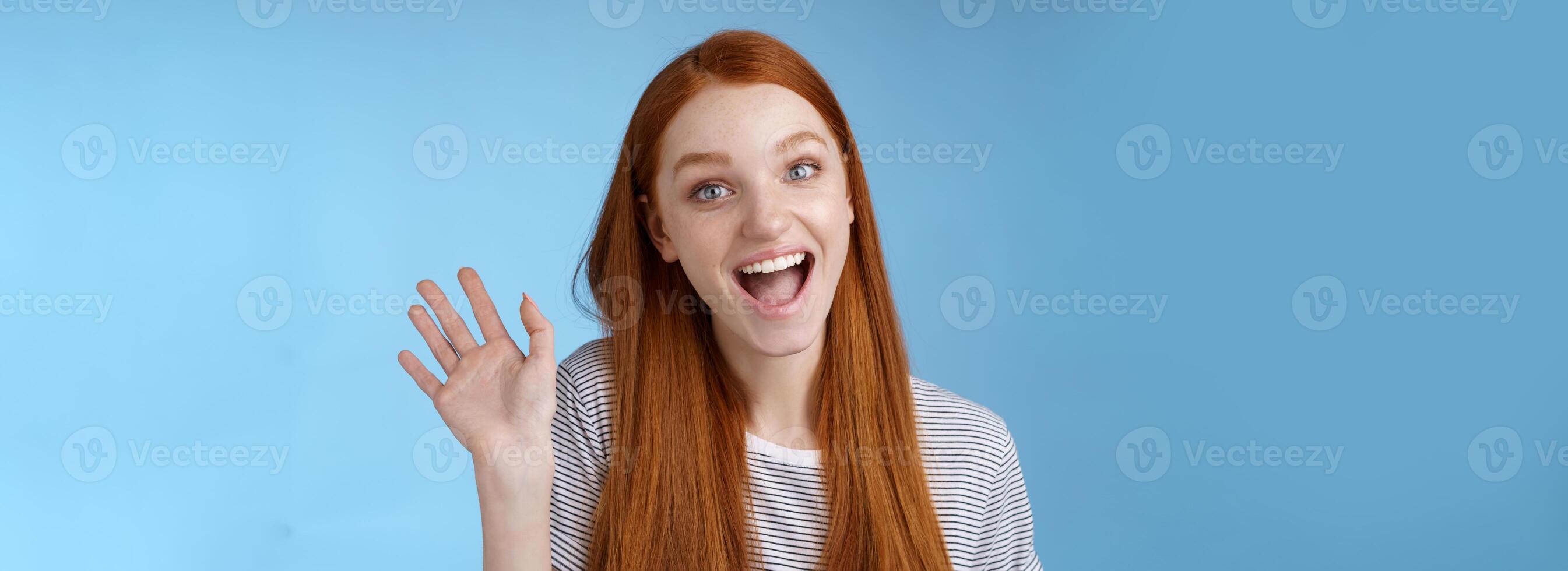 Hello wanna be friends. Enthusiastic cute redhead female newbie getting know coworkers smiling happy waving raised hand hi greeting gesture welcoming, say bye standing blue background photo