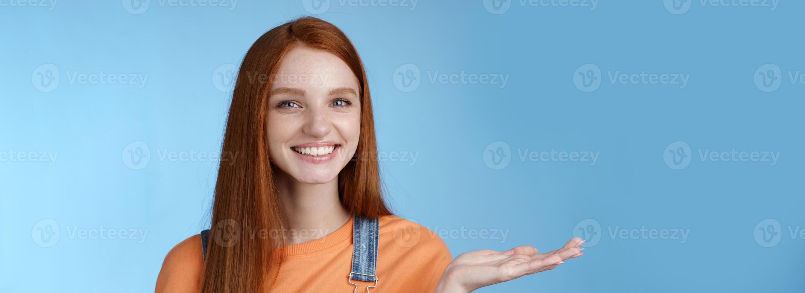 Proud good-looking confident redhead girl present awesome product hold object palm raise hand blank blue copy space smiling delighted recommend cool link, standing studio background helpful photo