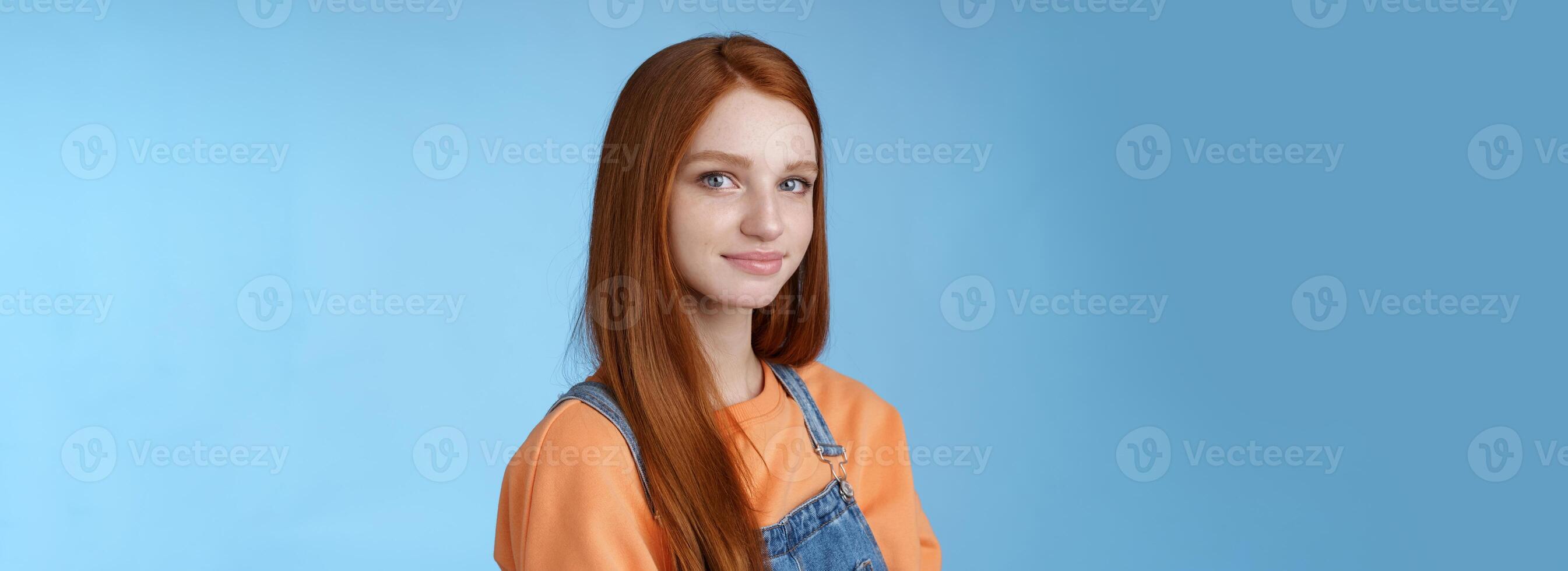 Waist-up kind sincere tender lovely redhead girl wearing orange shirt denim overalls standing half-turned smiling silly gentle grin camera looking friendly pleasantly walk alone blue background photo
