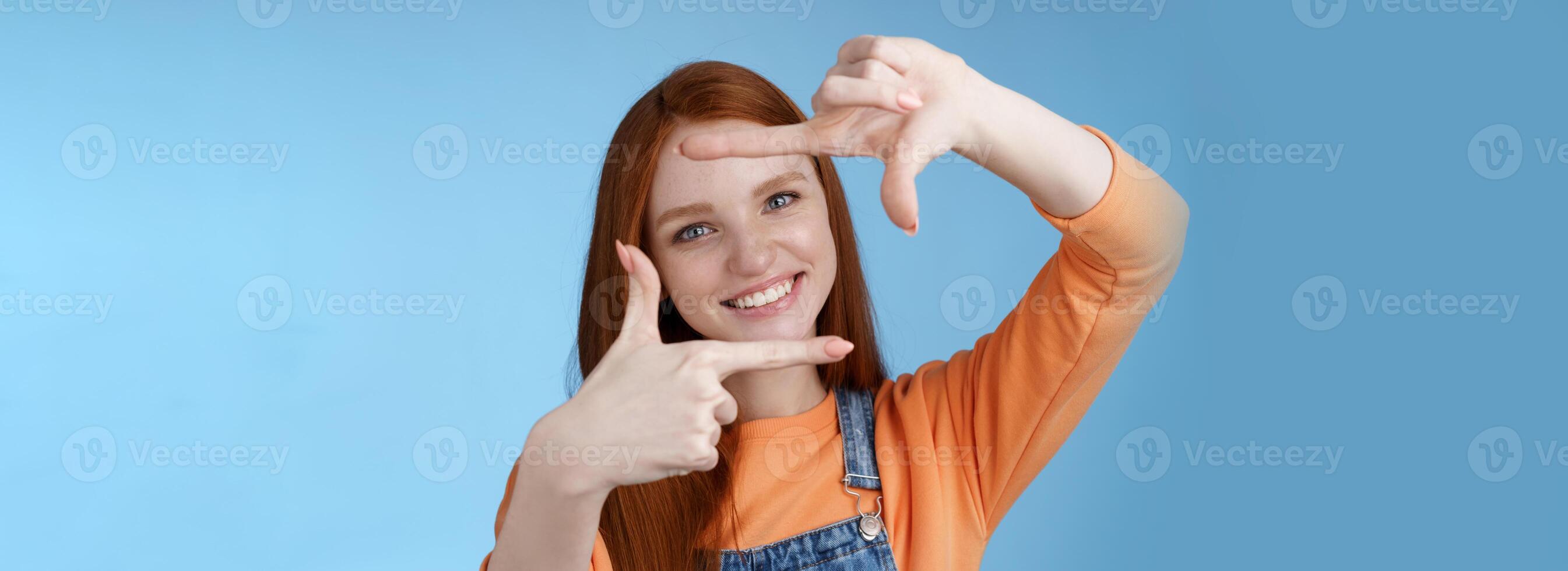 Joyful attractive sincere redhead young girl searching inspiration find perfect angle take good shot make hand frames look through delighted amused smiling broadly white teeth, blue background photo