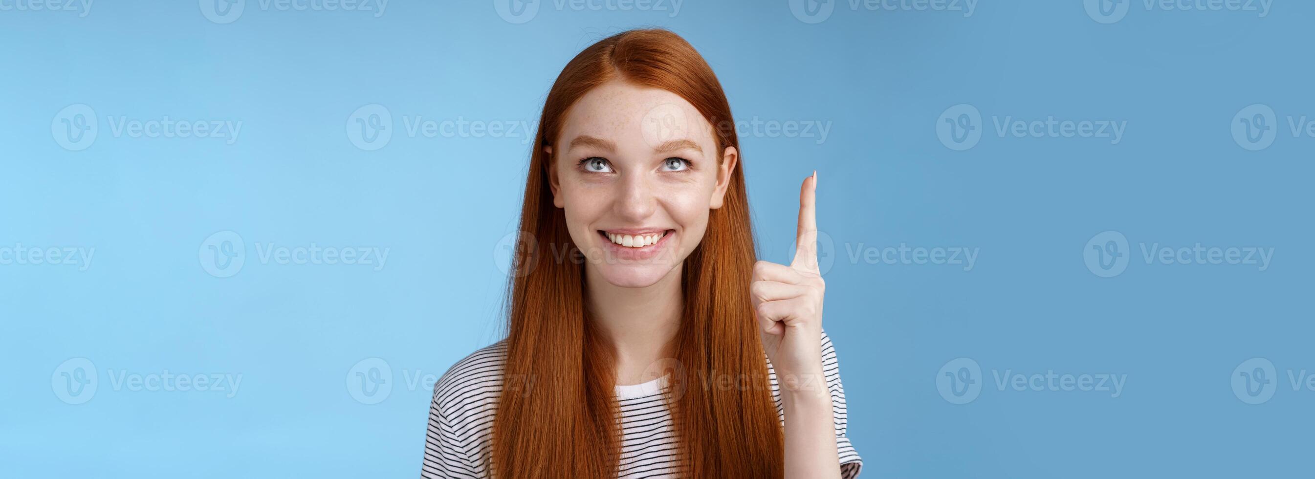 Intrigued lively happy smiling redhead european girl 20s look pointing up index fingers amused check out interesting sale holiday promo offer standing intrigued thrilled blue background photo