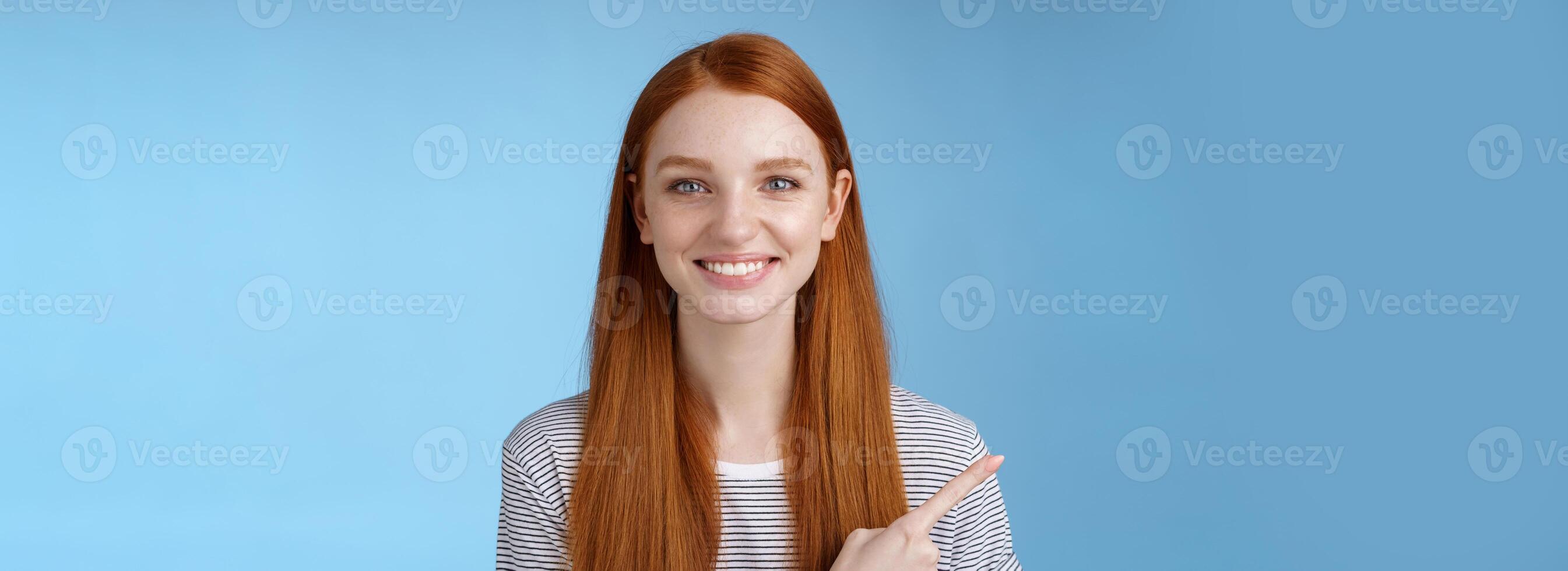 Pleasant charismatic helpful friendly-looking female redhead blue eyes pointing left index finger help make decision pick product store smiling broadly confident gaze camera asking trust her photo