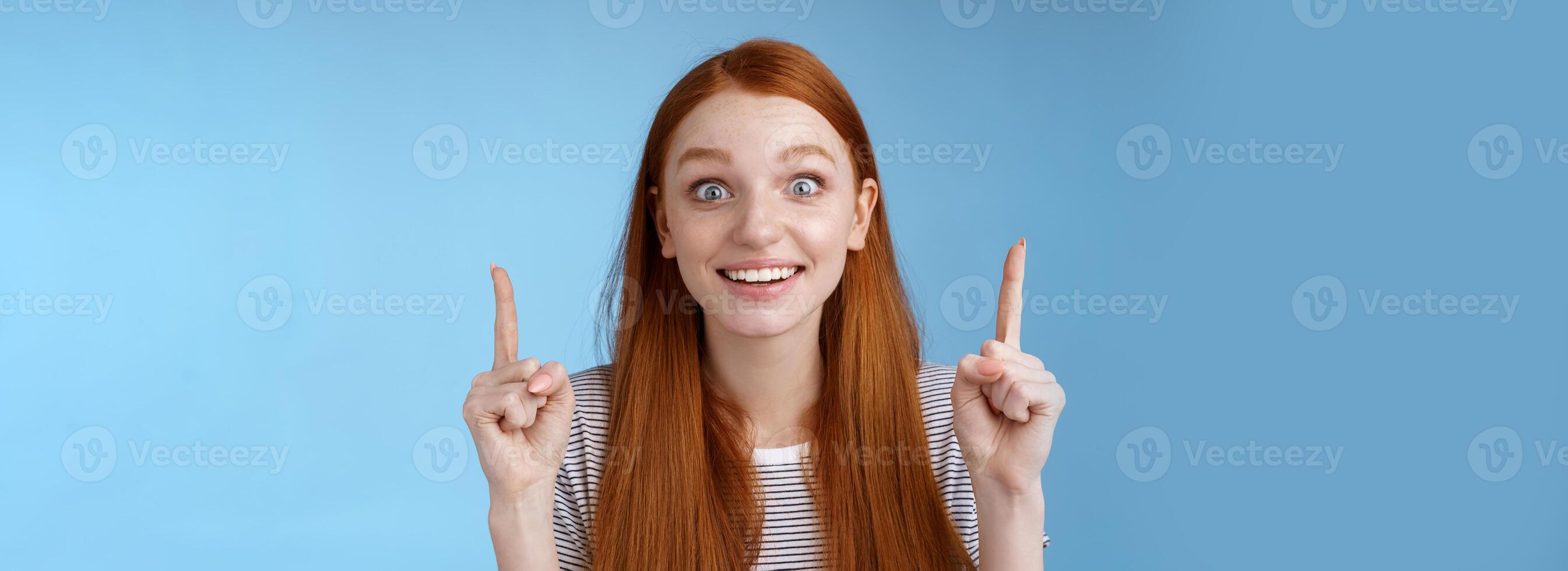 Thrilled happy young rejoicing redhead girl get awesome chance look surprised astonished smiling broadly wide eyes impressed incredible offer pointing up index fingers excitement, blue background photo