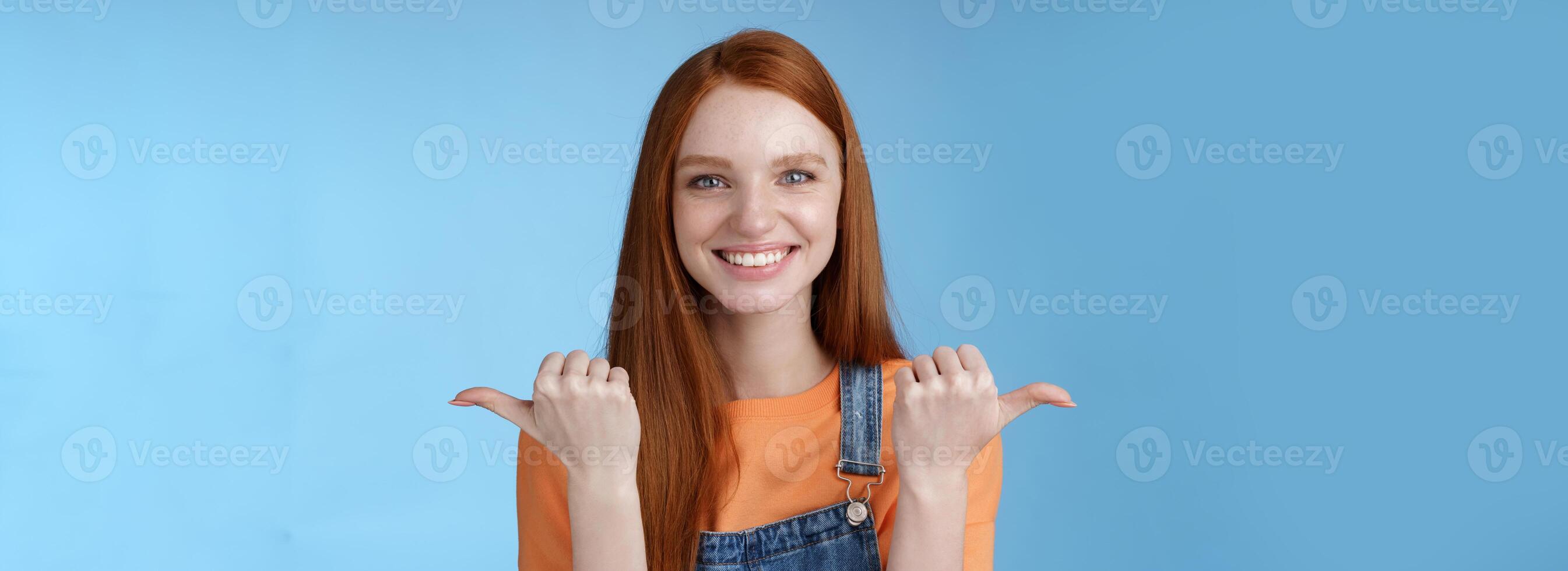 Indoor shot charismatic assertive happy smiling redhead woman orange shirt denim overalls pointing sideways thumbs left right showing choices opportunities give chance choose, blue background photo