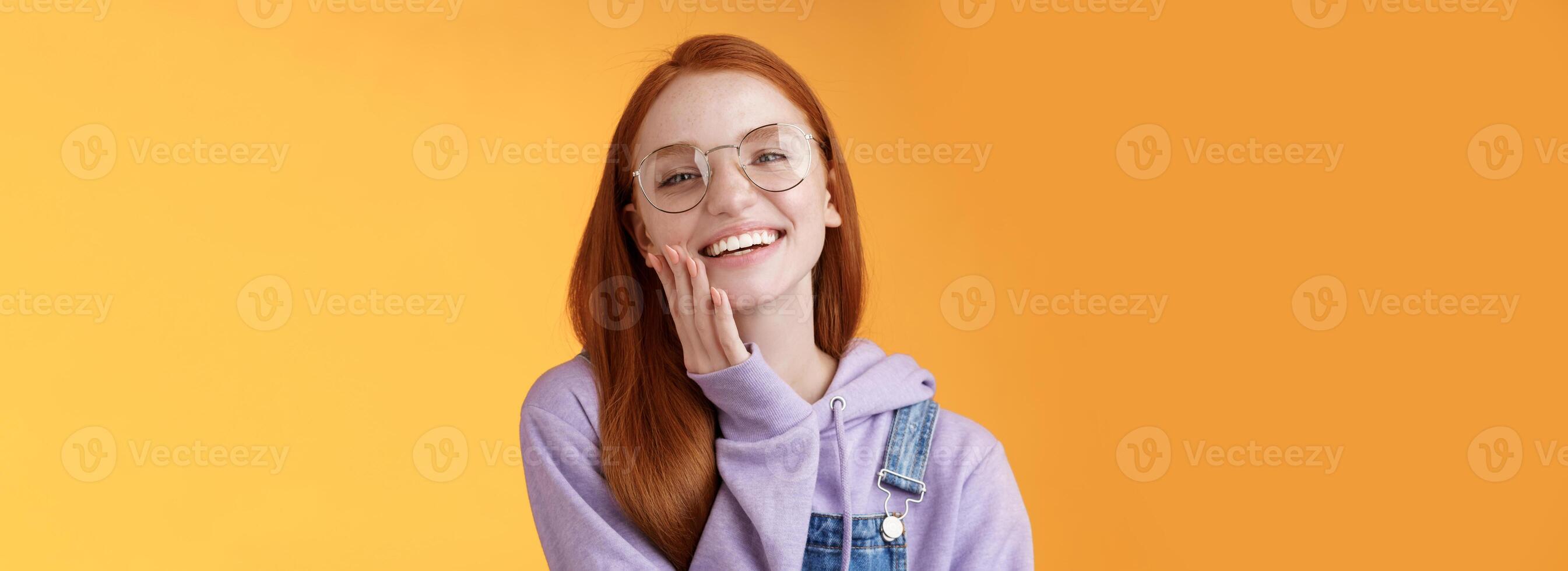 Charismatic happy tender redhead smiling european girl 20s glasses hoodie denim overalls having fun enjoying summer holidays chilly evening laughing joyfully touch pure clear skin, orange background photo