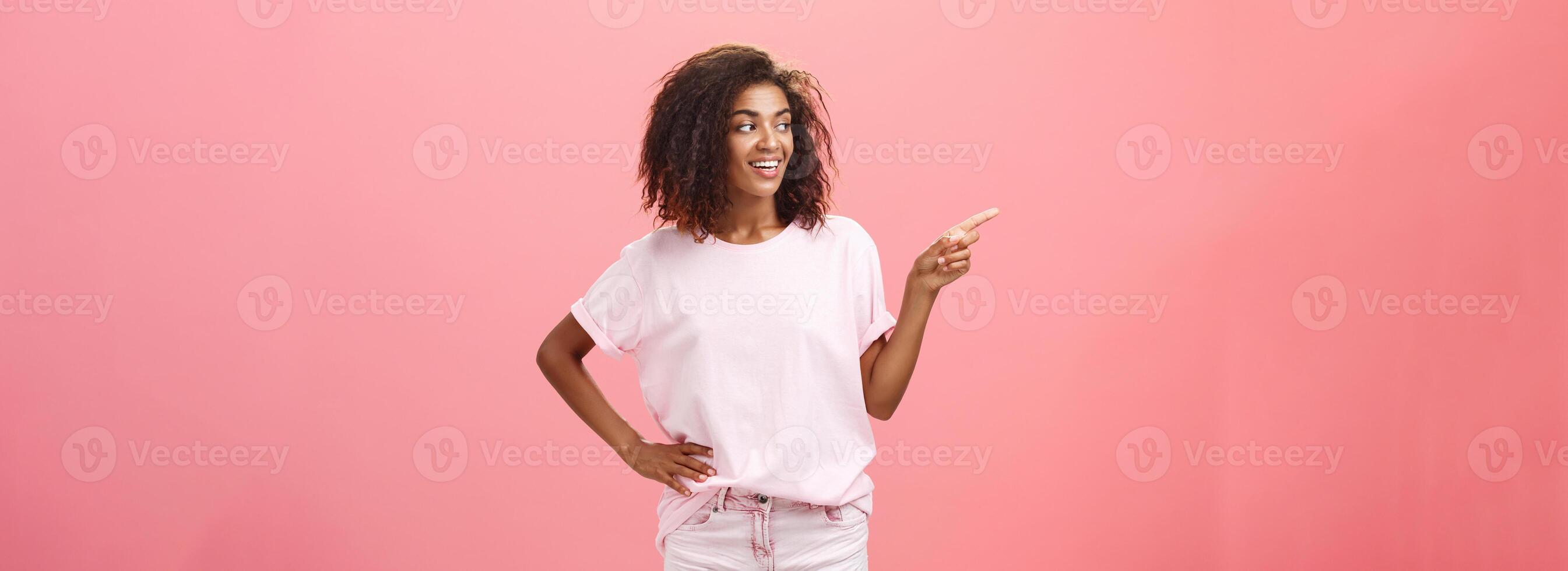 Girl checking out nice body of guy passing by. Charming confident dark-skinned female with curly hairstyle holding hand on hip looking and pointing left discussing cool copy space over pink wall photo
