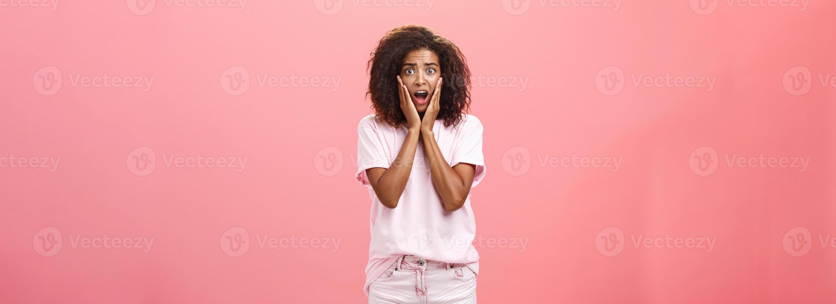 Worried, shocked african american female friend with curly hairstyle in trendy outfit being on vacation seeing scary animal shouting and gasping with opened mouth holding hands on face over pink wall photo