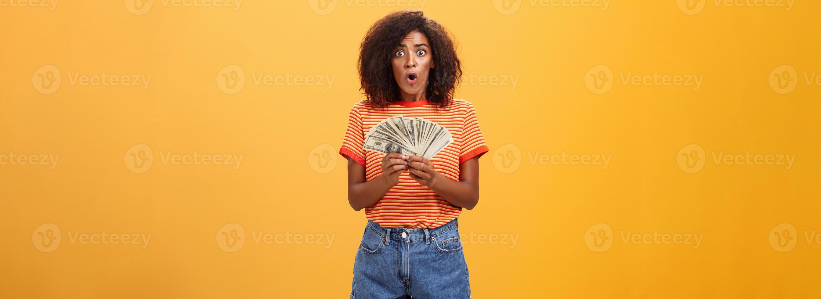 Woman shocked finding lots of cash in safe. Portrait of surprised speechless good-looking dark-skinned female with curly haircut folding lips gasping holding money posing over orange background photo