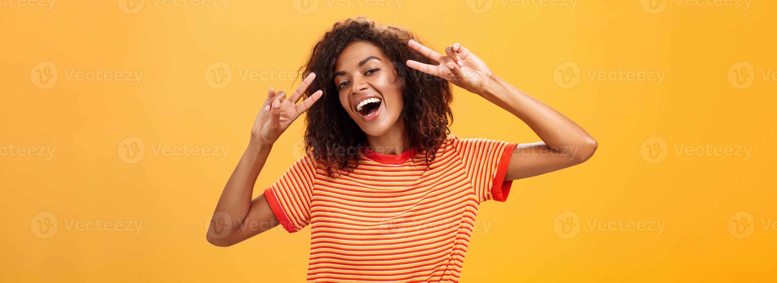 Nobody gonna spoil my perfect mood. Portrait of happy optimistic attractive dark-skinned female model with curly hairstyle tilting head smiling carefree showing peace or victory signs near face photo
