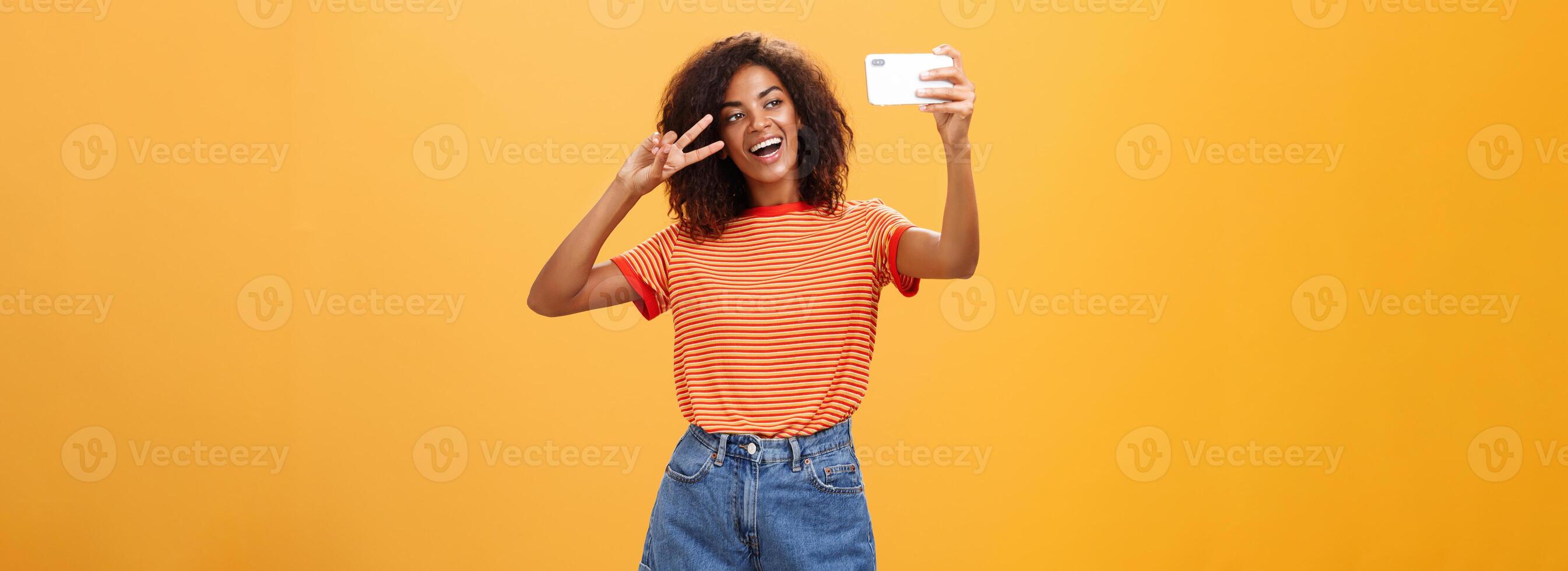 Girl making video vlog with brand new smartphone posting in internet trying become famous standing over orange background posing for selfie gazing at gadget screen showing peace or victory gesture photo